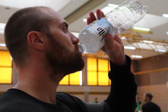 Ein Mann mit Bart trinkt Wasser aus einer Plastikflasche in einer Turnhalle mit gelb-orangenen Fenstern im Hintergrund.