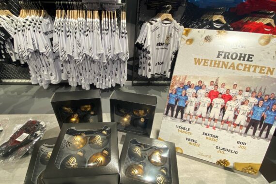 Auslage von Sporttrikots auf Ständern, Schachteln mit Goldschmuck auf einem Tisch und eine große Weihnachtskarte mit einer Sportmannschaft und Frohe Weihnachten auf Deutsch sowie anderen Weihnachtsgrüßen.