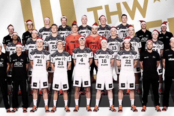 Eine Herren-Handballmannschaft posiert für ein Gruppenfoto in schwarz-weißen Trikots und roten Nikolausmützen. An den Seiten stehen vier Trainer oder Betreuer in schwarzen Outfits. Im Hintergrund ist ein großes goldenes Mannschaftslogo zu sehen.