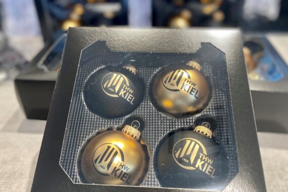 Eine schwarze Schachtel enthält vier Christbaumkugeln, die jeweils mit dem goldenen Logo und dem Text THW Kiel verziert sind. Die Ornamente sind in einem 2x2-Raster angeordnet.