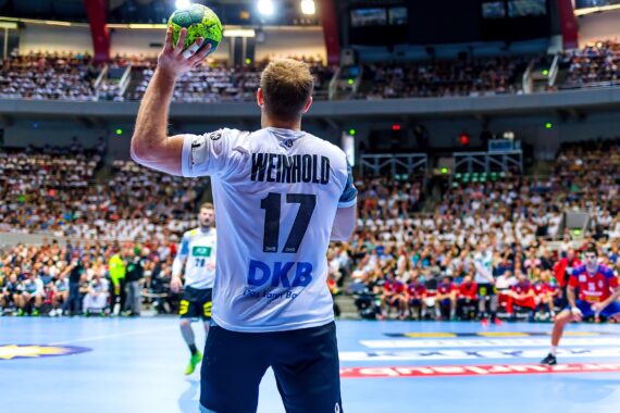 Ein Handballspieler, der ein weißes Trikot mit der Weinhold 17 trägt, bereitet sich während eines Spiels in einer vollbesetzten Halle auf den Wurf vor, wobei im Hintergrund Zuschauer und andere Spieler zu sehen sind.