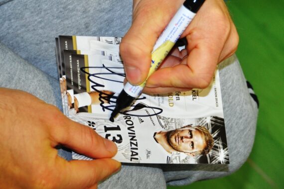 Eine Person signiert mit einem schwarzen Marker eine Sportkarte, auf der ein Sportler mit der Nummer 13 und dem Wort "PROVINZIAL" abgebildet ist. Das Porträt des Sportlers und ein Teil seines Trikots sind auf der Karte zu sehen.