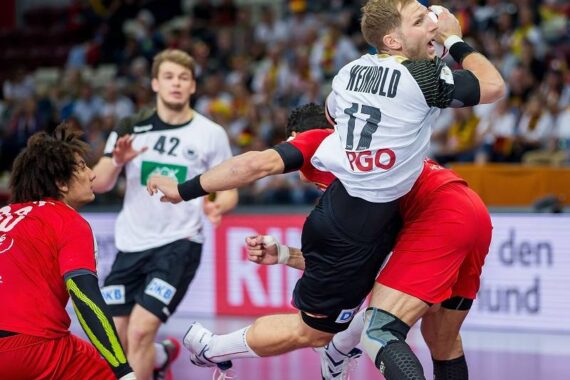 Ein Handballspieler in einem weißen Trikot springt, während er den Ball hält, der von einem Gegner in Rot eng verteidigt wird. Im Hintergrund sind andere Spieler und eine jubelnde Menge zu sehen.