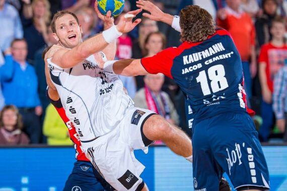 Ein Handballspieler in einem weißen Trikot springt, um den Ball zu schießen, während er von einem Spieler in einem blauen Trikot eng verteidigt wird. Im Hintergrund beobachten Zuschauer das intensive Spiel.