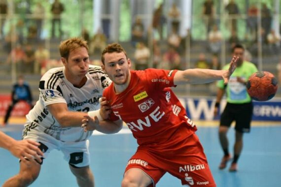 Zwei männliche Handballspieler kämpfen auf einem Hallenplatz um den Ball; der eine in einem roten Trikot hält den Ball, während er von einem Spieler in einem weißen Trikot eng verteidigt wird. Im Hintergrund sind Zuschauer und Offizielle zu sehen.