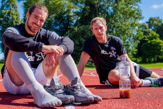 Zwei Männer in Sportkleidung sitzen und entspannen sich auf einer roten Laufbahn unter einem blauen Himmel. Beide lächeln, der eine sitzt im Schneidersitz, der andere mit ausgestreckten Beinen neben einer Wasserflasche. Im Hintergrund sind Bäume und Grünflächen zu sehen.
