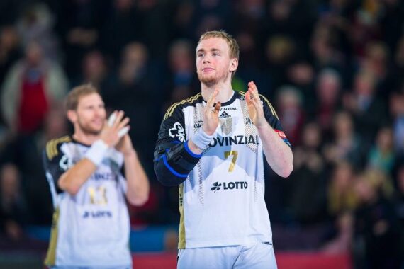 Zwei männliche Handballspieler in weißen Trikots mit schwarz-goldenen Akzenten applaudieren. Der vordere Spieler trägt die Nummer 7, und im Hintergrund ist eine unscharfe Menschenmenge zu sehen.