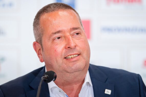Ein Mann mittleren Alters in Anzugjacke und weißem Hemd spricht auf einer Pressekonferenz in ein Mikrofon, wobei der Hintergrund mit Text und Logos unscharf ist.