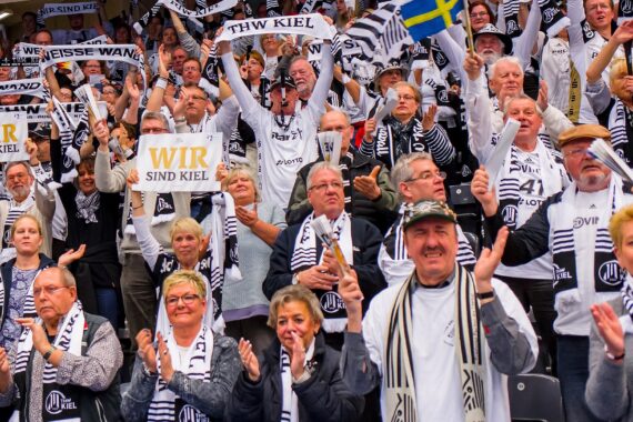 Eine große, begeisterte Menschenmenge in schwarz-weißer THW-Kleidung jubelt bei einer Sportveranstaltung und hält Schals und Schilder mit den Aufschriften WIR SIND KIEL und WEISSE WAND in die Höhe. Im Hintergrund ist eine schwedische Flagge zu sehen.