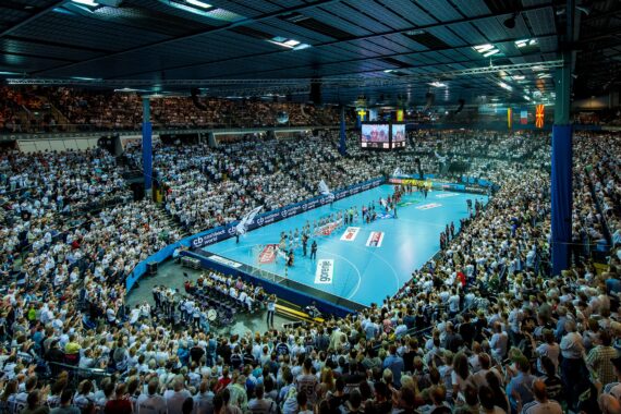 Eine große Sporthalle mit vielen Zuschauern, die ein Handballspiel verfolgen, mit Mannschaften, die auf einem blauen Spielfeld aufgereiht sind und hellen Lichtern, die die energiegeladene, überfüllte Szene beleuchten.
