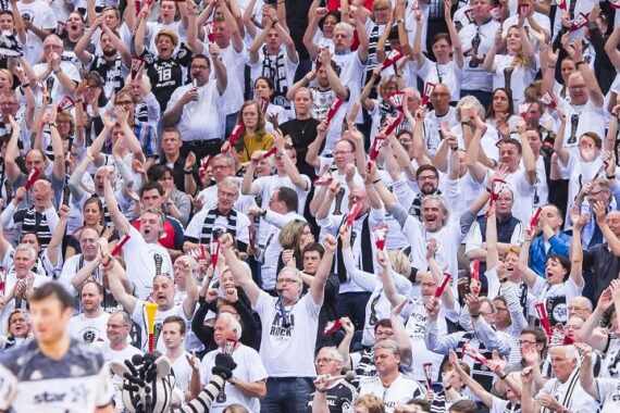 Eine große Menge begeisterter Sportfans in weißen Hemden jubelt und winkt mit ihren Schals, was eine lebhafte und energiegeladene Atmosphäre auf den Tribünen schafft.