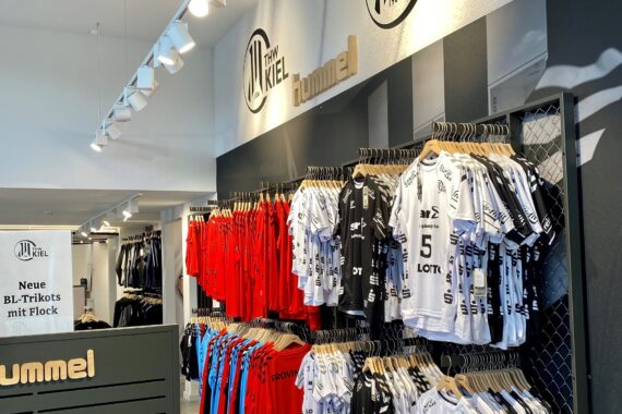 Eine Auslage mit roten, schwarzen und weißen Handballtrikots, die auf Regalen in einem Einzelhandelsgeschäft hängen, mit einem THW Kiel-Logo und einem Hummel-Schild über der Auslage. Auf einem Schild steht: Neue BL-Trikots mit Flock.