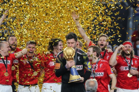 Eine Gruppe von Sportlern in roten Trikots feiert, jubelt und lächelt, während goldenes Konfetti um sie herum fällt. Ein Spieler steht in der Mitte und hält eine große goldene Trophäe in der Hand und küsst sie.