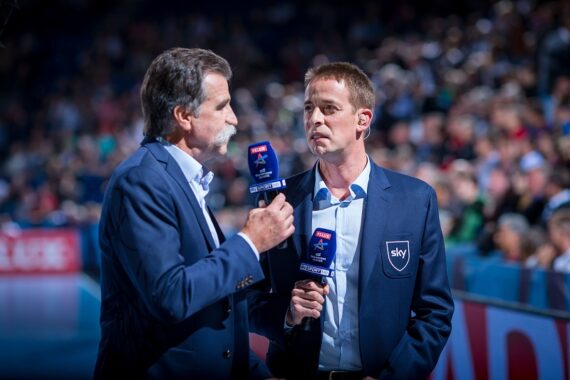 Zwei männliche Sportkommentatoren in Anzügen halten Mikrofone mit Sky-Logos in der Hand und stehen vor einer verschwommenen Menschenmenge bei einer Hallensportveranstaltung.