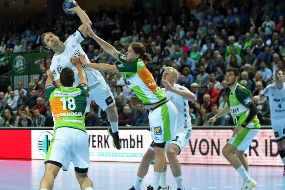 Ein Handballspieler in einem weißen Trikot springt, um den Ball zu schießen, während er von Verteidigern in grün-weißen Trikots bei einem überfüllten Hallenspiel geblockt wird. Die Zuschauer schauen von den Tribünen im Hintergrund zu.