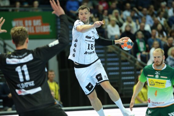 Ein Handballspieler in einem weißen Trikot springt, um den Ball zu werfen, während er von zwei Spielern in schwarzen und grünen Trikots während eines Spiels verteidigt wird, während die Zuschauer im Hintergrund zuschauen.