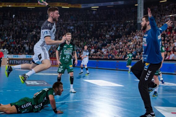 Ein Handballspieler in einem weiß-schwarzen Trikot springt in der Luft, um einen Schuss abzugeben, der auf das Tor zielt, während der Torwart sich darauf vorbereitet, ihn abzublocken. Ein anderer Spieler in grüner Kleidung liegt auf dem Spielfeld, und die Zuschauer auf der Tribüne schauen zu.