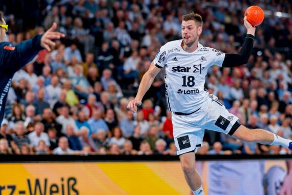 Ein männlicher Handballspieler in einem weißen Trikot mit der Nummer 18 springt in der Luft, um einen orangefarbenen Ball zu werfen, während er einem Verteidiger gegenübersteht. Die Zuschauer im Hintergrund verfolgen aufmerksam das Spiel.