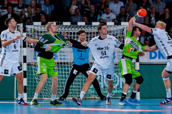 Fünf männliche Handballspieler, drei in weißen und zwei in grünen Trikots, liefern sich einen intensiven Wettkampf in Tornähe; ein weißer Spieler blockt einen Schuss der grünen Mannschaft ab, während der Torwart im Hintergrund wachsam zusieht.