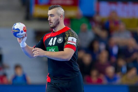 Ein männlicher Handballspieler in einem schwarz-roten Trikot mit der Nummer 48 hält einen Handball und bereitet sich darauf vor, ihn während eines Spiels zu werfen. Die Zuschauer sind im Hintergrund unscharf.