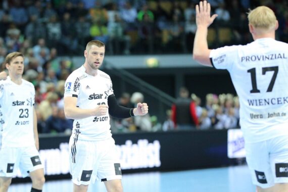 Drei männliche Handballspieler in weißen Trikots stehen auf einem Hallenplatz. Einer blickt mit erhobener Faust in die Kamera, während ein anderer, der die Nummer 17 trägt, mit dem Rücken zur Kamera steht. Der Hintergrund ist mit Zuschauern gefüllt.