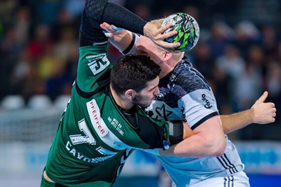 Zwei männliche Handballspieler in Aktion während eines Spiels; der eine im grünen Trikot versucht, den Ball zu blocken oder einem Gegner im grauen Trikot den Ball zu klauen, beide sind sehr konzentriert und körperlich engagiert.