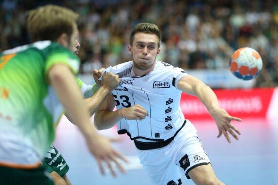 Ein männlicher Handballspieler in einem weißen Trikot wirft den Ball, während er von zwei Spielern in grünen Trikots während eines Hallenspiels verteidigt wird, mit einer verschwommenen Menschenmenge im Hintergrund.