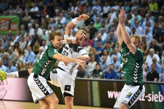 Drei männliche Handballspieler sind während eines Spiels in Aktion. Ein Spieler in einem weißen Trikot versucht einen Wurf, während zwei Spieler in grünen Trikots versuchen, ihn zu blocken. Eine Menschenmenge schaut von der Tribüne im Hintergrund zu.