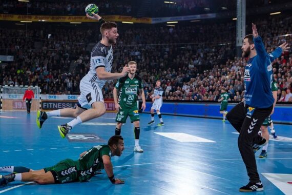 Ein Handballspieler in einem grau-weißen Trikot springt, um den Ball zu schießen, während ein grün gekleideter Verteidiger am Boden liegt und ein anderer Spieler und der Torwart sich darauf vorbereiten, den Ball zu blocken. Eine große Menschenmenge schaut von der Tribüne aus zu.
