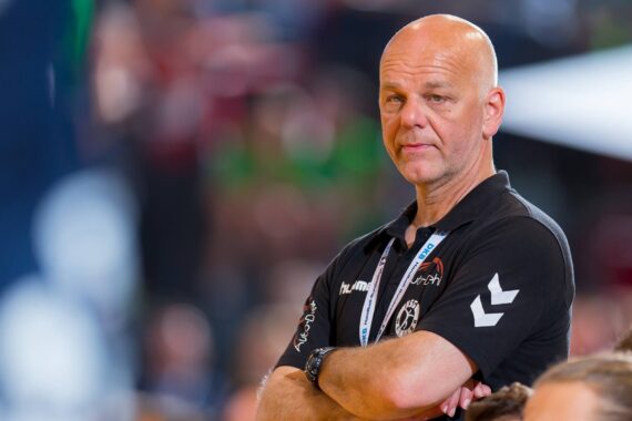 Ein glatzköpfiger Mann in einem schwarzen Polohemd mit weißen Logos steht mit verschränkten Armen da und blickt in einer hell erleuchteten Halle, möglicherweise bei einer Sportveranstaltung, zur Seite. Der Hintergrund ist durch Menschen und Lichter unscharf.