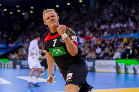 Ein männlicher Handballspieler in einem schwarzen Trikot mit der Nummer 7 ballt die Faust und jubelt ausgelassen auf dem Spielfeld, während im Hintergrund eine verschwommene Menschenmenge und andere Spieler zu sehen sind.