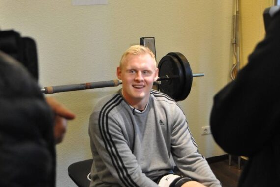 Ein lächelnder Mann mit blondem Haar in einem grauen Sweatshirt sitzt auf einer Hantelbank in einem Fitnessstudio, mit einer Langhantel und Gewichten hinter ihm. Zwei Personen sind teilweise im Vordergrund zu sehen.