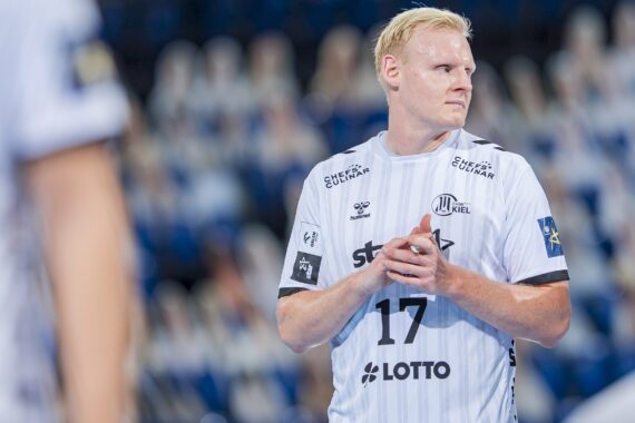 Ein männlicher Handballspieler in einem weißen Trikot mit der Nummer 17 steht auf einem Spielfeld und schaut mit gefalteten Händen zur Seite. Der Hintergrund ist mit blauen und weißen Farben verschwommen, was auf eine Halle hindeutet.