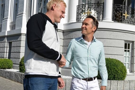 Zwei Männer stehen vor einem Gebäude, lächeln und schütteln sich die Hände. Ein Mann trägt eine schwarz-weiße Jacke mit Jeans, der andere ein hellgrün gestreiftes Hemd mit weißer Hose. Im Hintergrund sind weiße Säulen und ordentlich geschnittene Sträucher zu sehen.