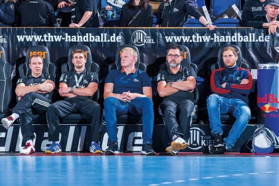 Fünf Männer sitzen auf einer Bank neben einem Handballfeld und beobachten das Spiel. Hinter ihnen ist ein Banner mit "thw-handball.de" zu sehen. Auf der rechten Seite des Bildes ist eine Red-Bull-Kühlbox zu sehen.