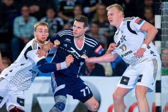 Drei männliche Handballspieler sind während eines Spiels in Aktion. Zwei Spieler in weißen Uniformen verteidigen gegen einen Spieler in einer dunkelblauen Uniform. Im Hintergrund ist eine unscharfe Zuschauermenge zu sehen.