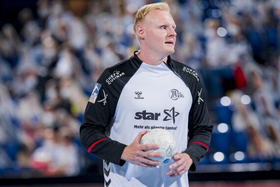 Ein männlicher Handballspieler in einem weiß-schwarzen Trikot, der einen Handball hält, steht auf einem Hallenplatz mit unscharfen Zuschauern im Hintergrund.