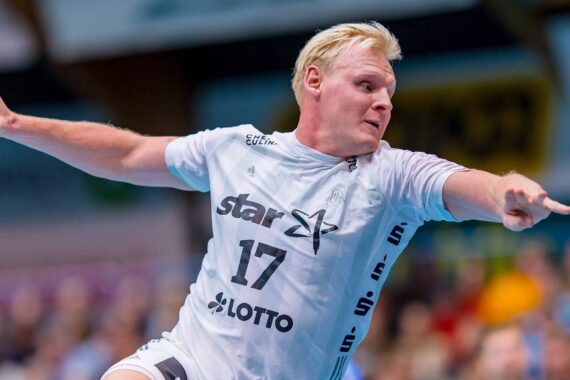 Ein männlicher Handballspieler in einem weißen Trikot mit der Nummer 17 befindet sich mitten in einer Aktion mit erhobenem Arm, hält einen Ball und scheint sich auf einen Wurf während eines Spiels vorzubereiten. Im Hintergrund ist eine unscharfe Menschenmenge zu sehen.