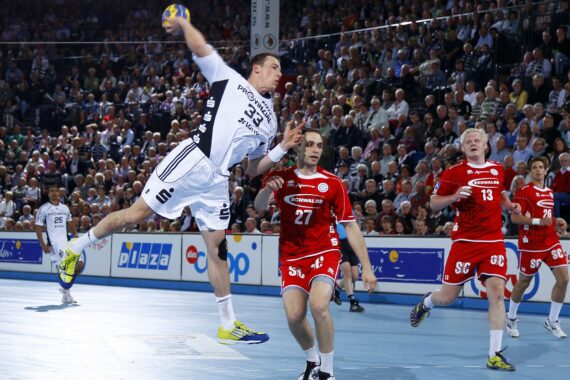 Ein Handballspieler in einem weißen Trikot springt, um den Ball zu schießen, während drei Spieler in roten Trikots den Ball verteidigen. Das Geschehen spielt sich in einer Halle ab, mit einer großen, jubelnden Menge im Hintergrund.