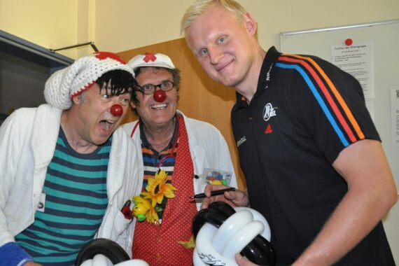 Ein lächelnder Mann in einem Sporthemd steht neben zwei Personen, die als Clowns mit roten Nasen und bunten Kostümen verkleidet sind. Die Gruppe wirkt fröhlich, und der Mann hält schwarze und weiße Ballonskulpturen in der Hand.