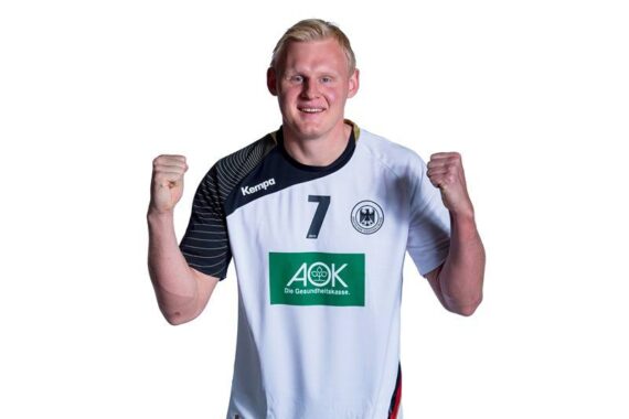 Ein männlicher Sportler in einem weißen Trikot mit der Nummer 7 und deutschen Symbolen steht lächelnd mit erhobenen Fäusten vor einem schlichten weißen Hintergrund.