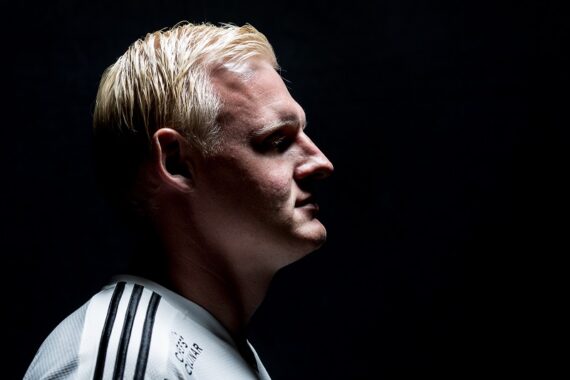Ein Mann mit kurzen blonden Haaren, der ein weißes Hemd mit schwarzen Streifen auf der Schulter trägt, ist im Profil vor einem dunklen Hintergrund dargestellt.