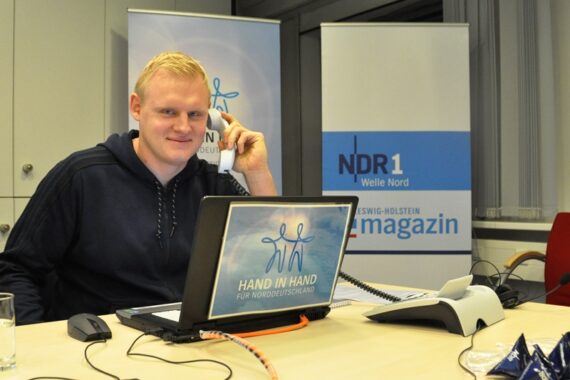 Ein Mann sitzt an einem Schreibtisch, lächelt und hält ein Telefon an sein Ohr. Vor ihm steht ein Laptop, auf dem der Text Hand in Hand für Norddeutschland steht. Im Hintergrund sind Banner von NDR 1 und dem Schleswig-Holstein Magazin zu sehen.
