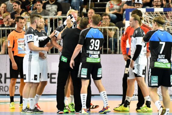 Eine Gruppe männlicher Handballspieler und ein Trainer versammeln sich auf dem Spielfeld, wobei ein Spieler wegen seines Gesichts medizinisch behandelt wird. Die Zuschauer schauen von der Tribüne im Hintergrund zu.