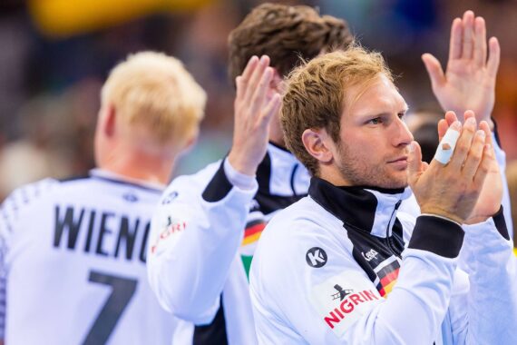 Mehrere männliche Sportler in weißen Trikots mit schwarzen und grauen Details klatschen in die Hände und schauen in verschiedene Richtungen. Auf dem Trikot eines Spielers steht der Name WIENEKE und die Nummer 7. Sie scheinen die Menge zu grüßen.