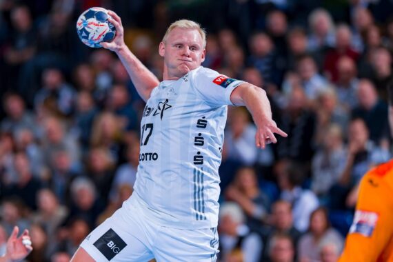 Ein Handballspieler in einem weißen Trikot bereitet sich darauf vor, den Ball während eines Spiels in die Luft zu werfen, während ein Spieler in einem orangefarbenen Trikot vor ihm steht. Im unscharfen Hintergrund sieht die Menge zu.