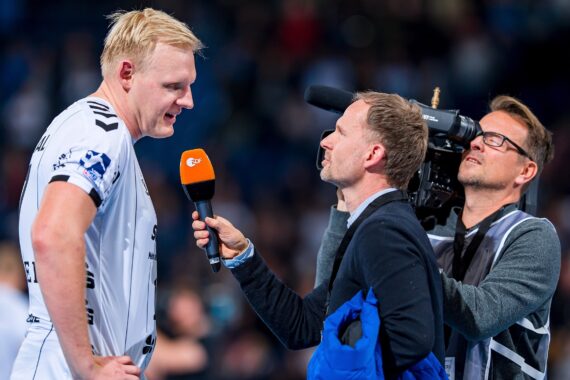 Ein männlicher Sportler in einem weißen Trikot wird von einem Reporter interviewt, der ein orangefarbenes Mikrofon hält, während ein Kameramann die Szene aufnimmt. Das Interview findet in einem Innenraum statt, möglicherweise nach einem Sportereignis.