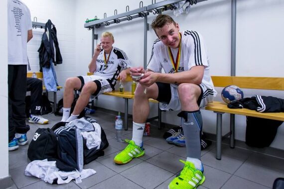Zwei männliche Sportler in einer Umkleidekabine in Sportkleidung sitzen auf Bänken, einer lächelt in die Kamera, der andere telefoniert. Sporttaschen, Kleidung und ein Handball liegen verstreut auf dem Boden.
