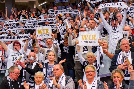 Eine große Menge begeisterter Sportfans, viele in Schwarz und Weiß, jubeln und halten Schals und Schilder mit der Aufschrift WEISSE WAND und WIR SIND KIEL auf den Tribünen einer Reithalle hoch.