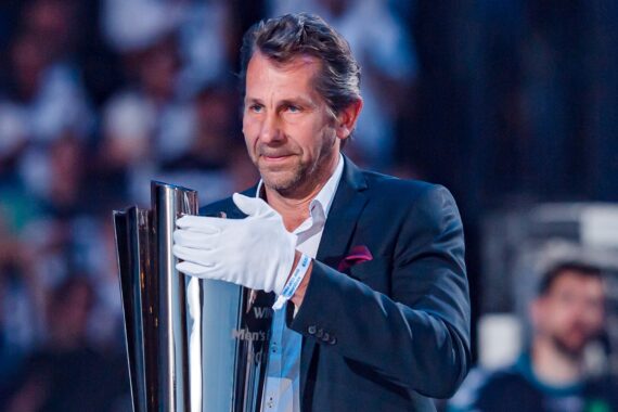 Ein Mann im Anzug und mit weißen Handschuhen hält eine große Trophäe. Er steht in einem Innenraum, im Hintergrund ist eine unscharfe Menschenmenge zu sehen.